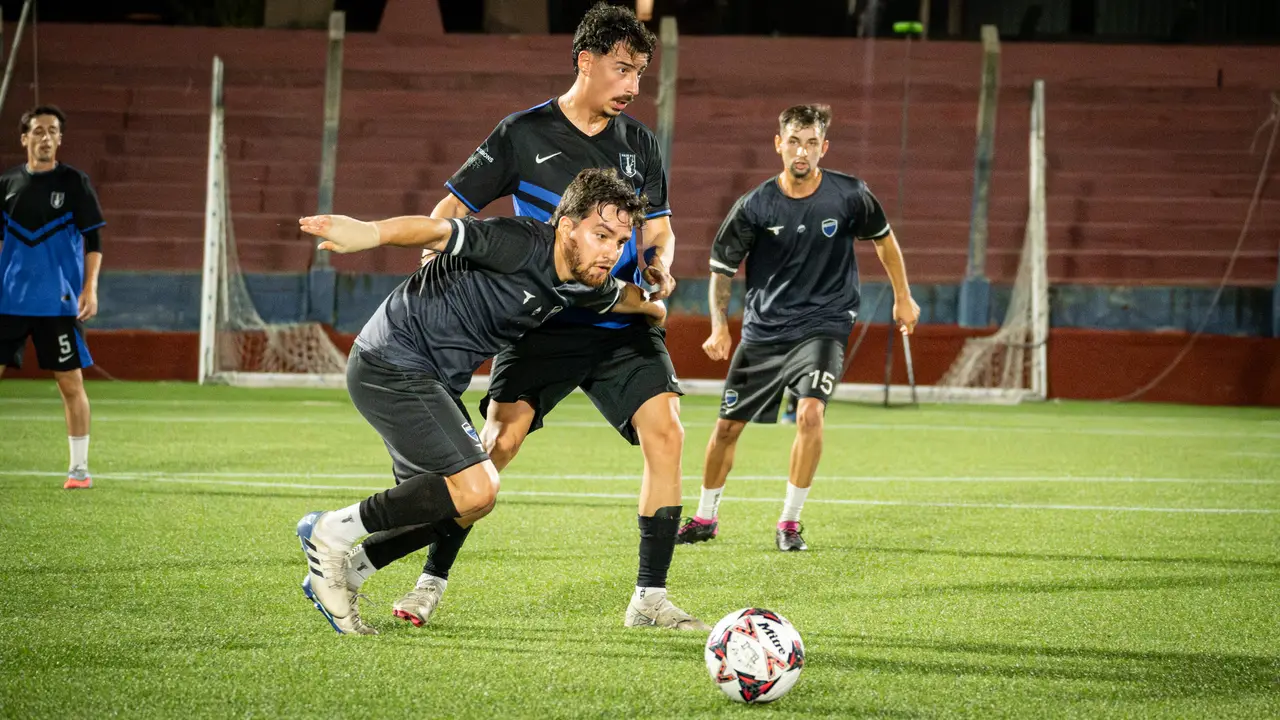 Jugador driblando entre rivales en partido nocturno con tribunas de fondo
