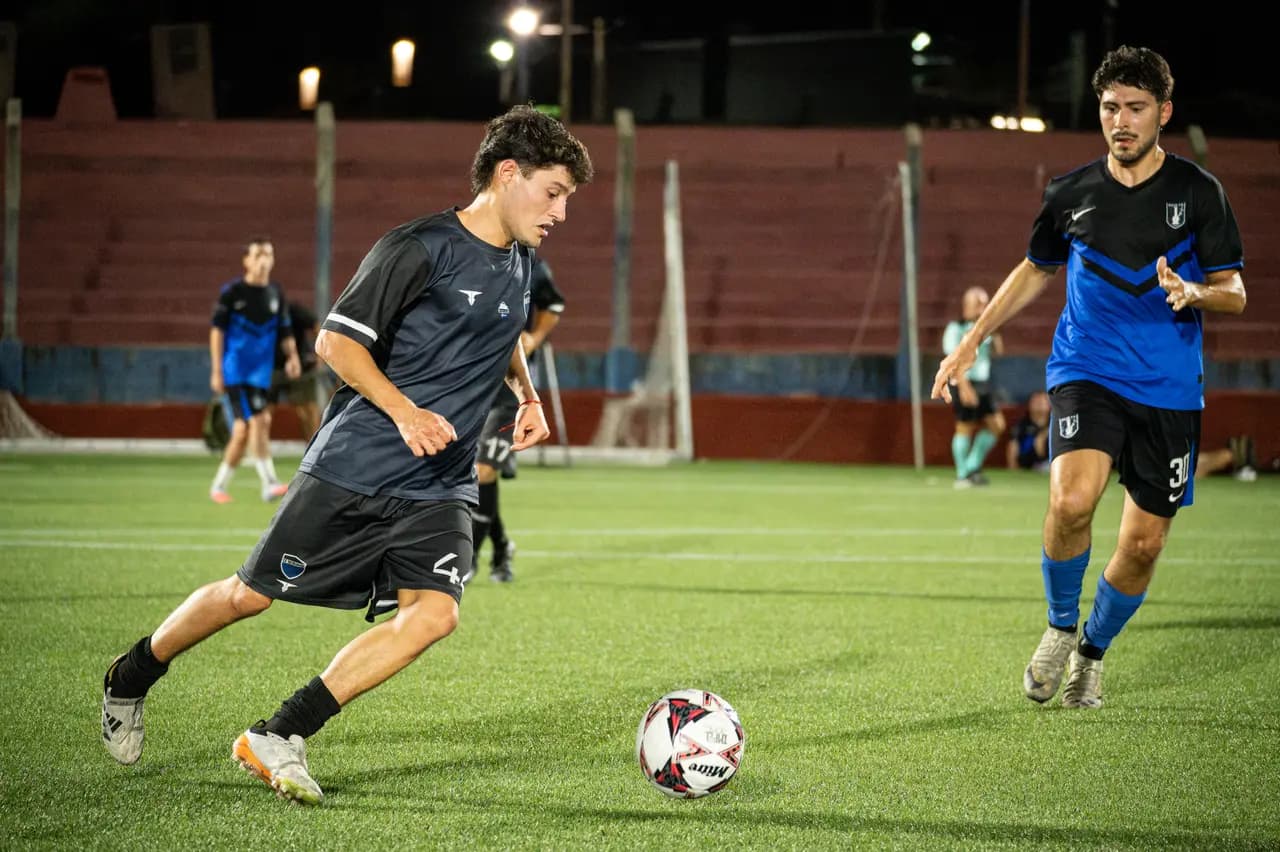 Jugador avanzando con la pelota en partido nocturno ante rival de camiseta azul